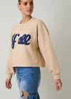 Woman wearing a beige sweatshirt with blue text and blue jeans on a plain background