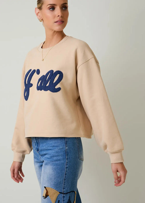 Woman wearing a beige sweatshirt with blue text and blue jeans on a plain background