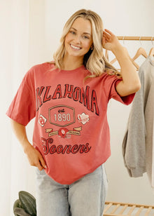 Woman wearing a red 'Oklahoma Sooners' t-shirt in a room with a white wall and wooden rack.