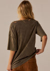 A woman shown from the back wearing a washed olive-brown short-sleeve top and brown pants against a plain light beige studio background.