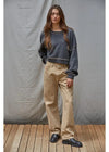 Woman wearing a dark blue sweater and beige pants standing against a gray background.
