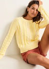 Woman wearing a yellow cable knit sweater and pink shorts sitting on a white surface.