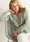 Woman wearing a light green embroidered jacket sitting on a chair with a white background