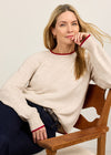 A woman sits on a wooden chair wearing a cream-colored knit sweater with red ribbed trim at the collar and cuffs, paired with dark navy pants, against a plain light background.