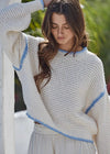 Woman wearing a white sweater with blue trim against a blurred outdoor background