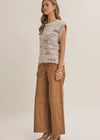 A woman modeling a sleeveless knit top with a textured, frayed hem in a light beige and brown stripe pattern, paired with wide-leg tan pants and beige heels, against a plain beige studio background.