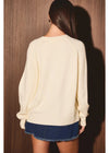 A woman shown from the back wearing a cream-colored knit sweater and a denim mini skirt, standing against a dark wood-paneled wall.