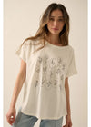 A woman modeling a cream-colored short-sleeve t-shirt with a black line-art graphic of various flowers arranged in a grid on the front, paired with light-wash blue jeans against a solid beige background.