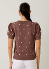 Woman wearing a brown sweater with white polka dots and white pants on a plain background