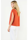 A woman with long blonde hair is shown from the back wearing a sleeveless, textured orange knit top and white pants against a plain white background.