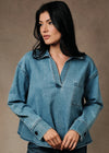 A woman wearing a light blue denim long-sleeve shirt with a half-placket and chest pocket, paired with dark blue jeans, set against a textured brown studio background.
