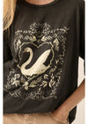 A close-up of a person wearing a black oversized T-shirt with a large white swan and ornate leafy border graphic on the front, paired with light beige pants.