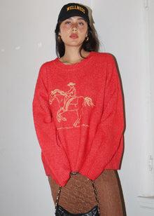  Person wearing a red sweater with a horse graphic and a black cap, standing against a white wall.