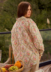 Woman wearing a floral jacket standing outdoors with a basket of fruits on a table.