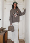 Woman wearing a sweater with bird design and holding a handbag in a room.