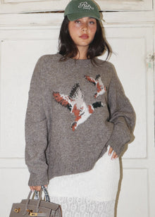  Person wearing a gray sweater with a fox design, holding a handbag indoors.