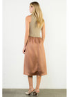 A woman stands with her back to the camera wearing a sleeveless two-piece outfit featuring a beige knit top and a brown satin midi skirt, paired with beige high-heeled sandals, set against a plain white wall.