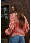 Woman wearing a red and white striped sweater and blue jeans standing against a wooden wall.