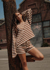 A woman posing outdoors in a brown and cream striped lounge set consisting of a long-sleeve top and matching shorts, leaning against a wooden utility pole in front of a brick building.