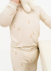 Child wearing a beige long-sleeve shirt and pants with duck pattern, holding white mittens.