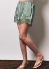 A model wearing a sage green satin mini skirt with a lace hem, paired with white heeled thong sandals, standing against a plain light gray wall.