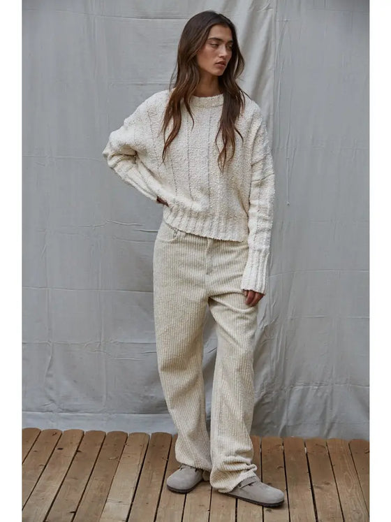 Woman wearing a white sweater and beige pants standing against a gray curtain.