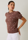 Woman wearing a brown sweater with white polka dots and white pants on a plain background