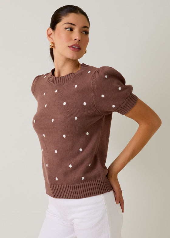Woman wearing a brown sweater with white polka dots and white pants on a plain background
