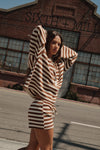 A woman wearing a matching brown and cream striped lounge set stands on a street in front of a brick industrial building with 'SIXTH MIL' visible on the facade.