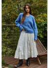 Woman wearing a blue sweater and white skirt standing outdoors with greenery in the background