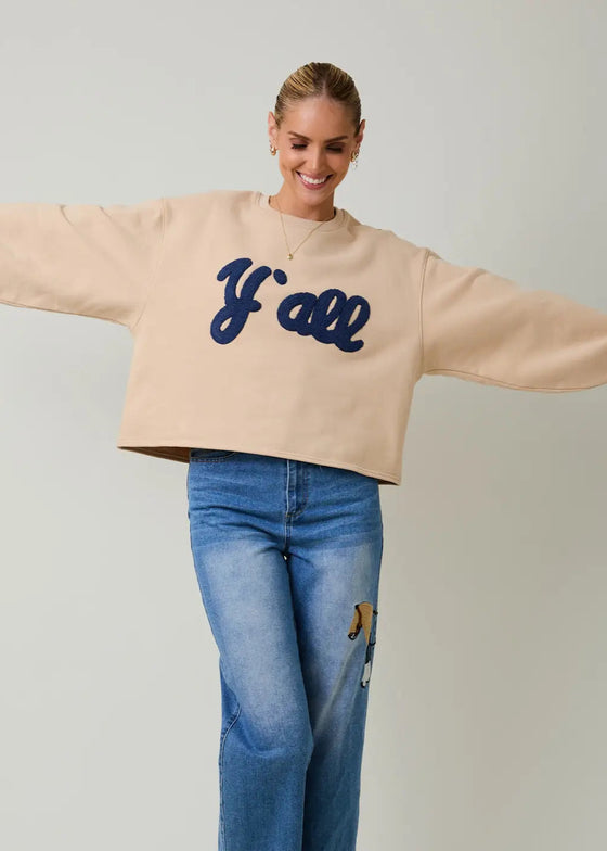 Person wearing a beige sweatshirt with 'Y'all' text and blue jeans on a plain background
