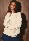 A woman wearing a cream-colored knit button-up henley sweater and a blue denim skirt, posing against a dark wood-paneled wall with dramatic lighting.