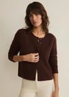 A woman wearing a dark brown button-front cardigan sweater and cream pants, standing against a plain light beige studio background.