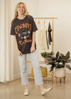 Woman wearing a 'Cowboys to Paris' t-shirt in a room with a clothing rack and plants.
