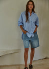 Woman wearing a blue striped shirt and denim shorts standing against a plain wall.