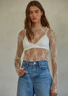 Woman wearing a sheer lace top over a white crop top and high-waisted jeans against a plain background