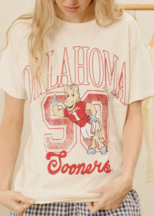  Person wearing a 'Oklahoma Sooners' t-shirt with a cartoon character on a beige background