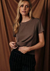 A woman modeling a short-sleeve crewneck t-shirt in a muted brown color, paired with black pinstriped trousers, set against a draped brown fabric backdrop.