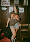 Woman wearing a 'POKES' t-shirt in a room with wooden walls and framed pictures.