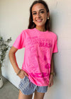 A smiling young woman wearing a bright pink graphic t-shirt with 'MONSTER MASH' printed on the front, paired with blue and white striped shorts, posing against a textured white wall.