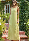 Woman wearing a light green pleated dress standing outdoors with greenery and a building in the background.