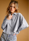 A woman wearing a light blue and white vertically striped button-up shirt with voluminous balloon sleeves, paired with light-wash jeans, posed against a solid warm brown background.
