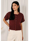 A woman modeling a short-sleeve burgundy knit cardigan with a button-up front, paired with white pants, set against a soft white curtain background.