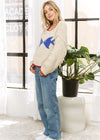 Woman wearing a cream sweater with a blue graphic design, standing indoors near a window.