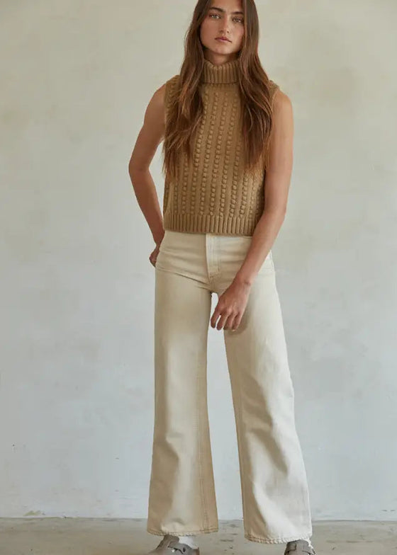 Woman wearing a brown sleeveless knitted top and beige pants against a plain background