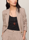 A woman wearing a beige pinstripe blazer and matching trousers over a black top, accessorized with a long pendant necklace and gold hoop earrings, against a light gray studio background.