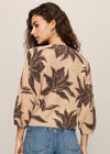 Woman wearing a beige sweater with brown leaf pattern and blue jeans on a plain background