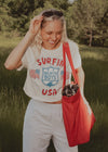 Woman wearing a 'Surfing Beach Boys USA' t-shirt with a red bag containing a small dog in a field.