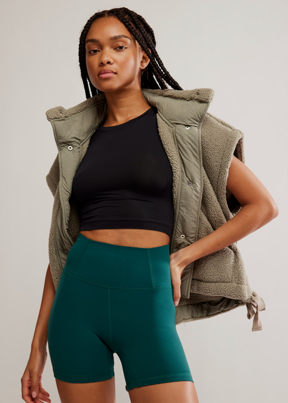 Woman wearing a black crop top and teal shorts with a green vest on a plain background
