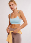 A woman modeling a light blue sports bra and taupe leggings, with a yellow towel tied around her waist, against a plain light gray studio background.